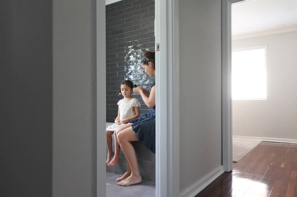 Mother braiding her daughters hair in bathroom. Domestic family life concept. Real people. Copy space - Foto, Imagem