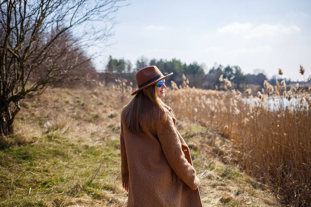 Ένα κορίτσι με καφέ παλτό, καπέλο και γυαλιά περπατάει σε ένα πάρκο με μια λίμνη κάτω από τον ήλιο. Απολαμβάνει τη ζωή και τα χαμόγελα. Η αρχή της άνοιξης - Φωτογραφία, εικόνα