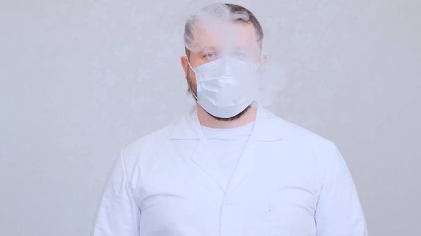 fehér ruhás férfi elektromos cigarettát szív egy orvosi maszkon keresztül. Vírusok, koronavírusok, tüdőbetegségek elleni védelem - Fotó, kép