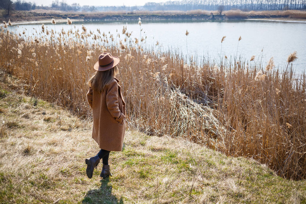 Egy barna kabátos, kalapos és szemüveges lány sétál a parkban egy tóval a fényes nap alatt. Örül az életnek és mosolyog. A tavasz kezdete - Fotó, kép