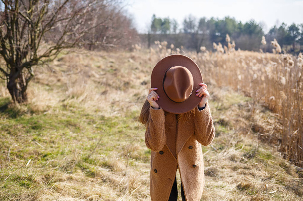 Egy barna kabátos, kalapos és szemüveges lány sétál a parkban egy tóval a fényes nap alatt. Örül az életnek és mosolyog. A tavasz kezdete - Fotó, kép