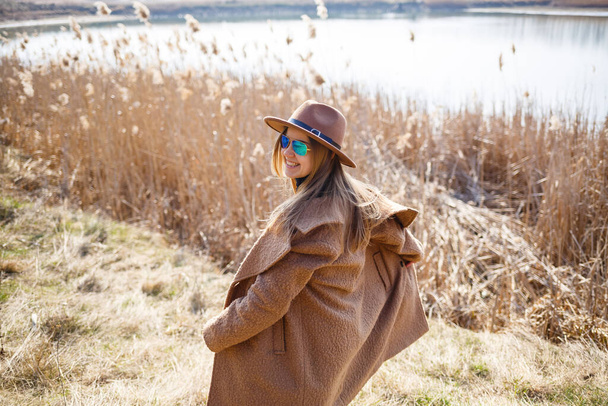 A girl in a brown coat, hat and glasses walks in a park with a lake under the bright sun. Rejoices in life and smiles. The beginning of spring - Photo, Image