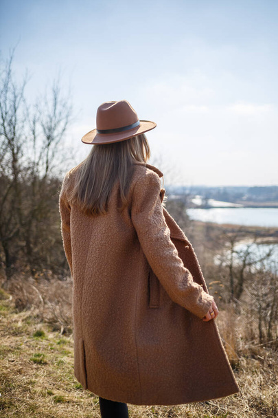 Une fille en manteau marron, chapeau et lunettes marche dans le parc avec un lac sous le soleil éclatant
. - Photo, image