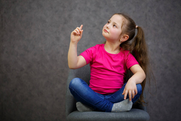 little girl in a pink t-shirt is sitting on a gray chair. Emotional photo of a child - 写真・画像