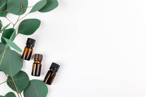 Unbranded amber glass bottles lying with green eucalyptus leaves. Cosmetic containers for natural medicine, essential and massage oil, aromatherapy products on white. Copy space in right side. - Fotografie, Obrázek