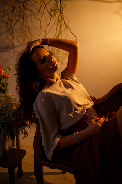 Una chica con iluminación marrón se sienta en un sofá. Alrededor de flores frescas en un jarrón
. - Foto, Imagen