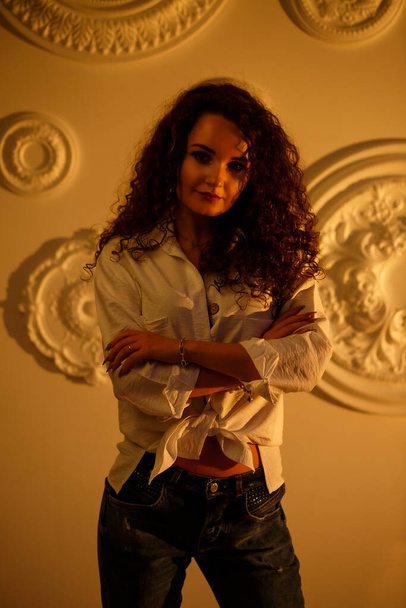 Krásná mladá dívka modelka s kudrlinkami pózování. Má na sobě bílou košili a džíny.. - Fotografie, Obrázek