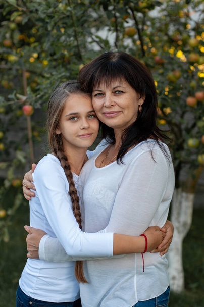 όμορφη μητέρα και η έφηβη κόρη της στέκονται σε μια αγκαλιά στη μέση ενός οπωρώνα μήλου - Φωτογραφία, εικόνα