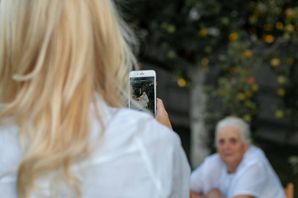 junges Mädchen mit langen blonden Haaren fotografiert ihre geliebte Großmutter im Garten - Foto, Bild