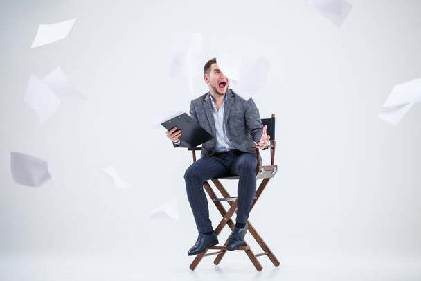 Επιχειρηματίας επιχειρηματίας με κοστούμι κάθεται σε μια ξύλινη καρέκλα και πετάει έγγραφα. Σε λευκό φόντο με σκέψεις για ένα νέο έργο - Φωτογραφία, εικόνα