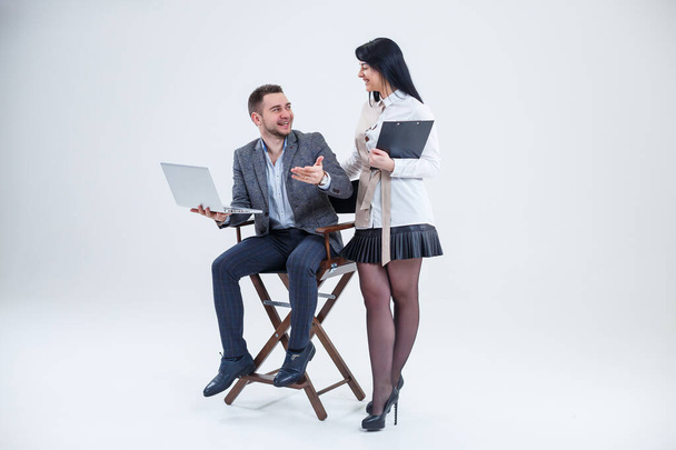A successful man in a suit is sitting on a black chair with a laptop and a woman is standing nearby. Future businessmen are smiling and talking about a new project. - Foto, imagen