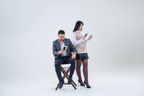 A successful man in a suit sits on a black chair and a woman stands next to look at the tablets. Future businessmen are smiling and talking about a new project. - Foto, immagini