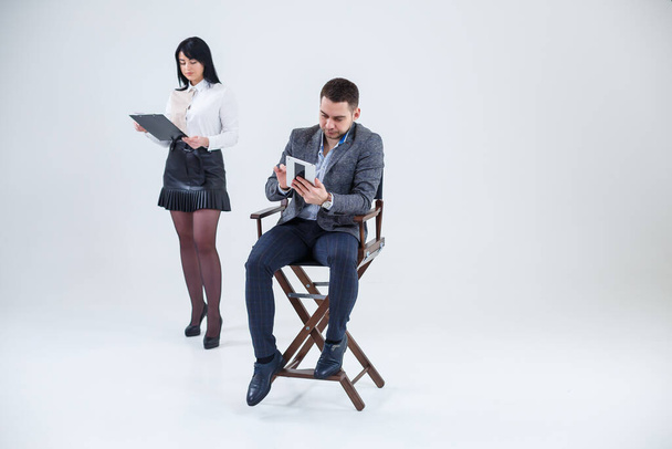 A successful man in a suit sits on a black chair with a tablet and a woman stands nearby looks in a folder. Future businessmen are smiling and talking about a new project. - Fotoğraf, Görsel