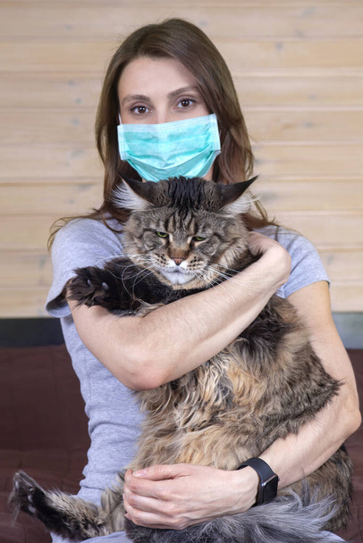 Young attractive woman with medical mask holds stunning cat of maine coon breed, with strong face expression. Virus and protective measures at home concept, safety for animals. Soft selective focus. - Photo, Image