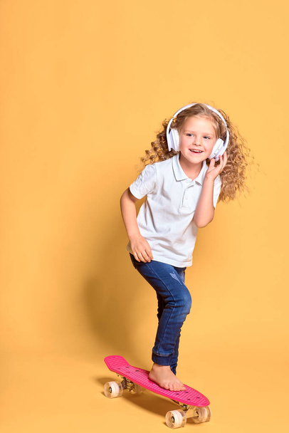 Active and happy girl with curly hair, headphones having fun with penny board, smiling face stand skateboard. Penny board cute skateboard for girls. Lets ride. Girl ride penny board yellow background. - Fotó, kép