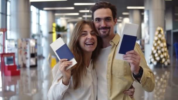 Ενθουσιασμένο ζευγάρι Καυκάσιων που πηγαίνουν διακοπές και κρατούν κάρτα επιβίβασης στο αεροδρόμιο. Κουνώντας με check-in διαβατήρια, χαμογελώντας στην κάμερα, αγκαλιάζοντας - Πλάνα, βίντεο