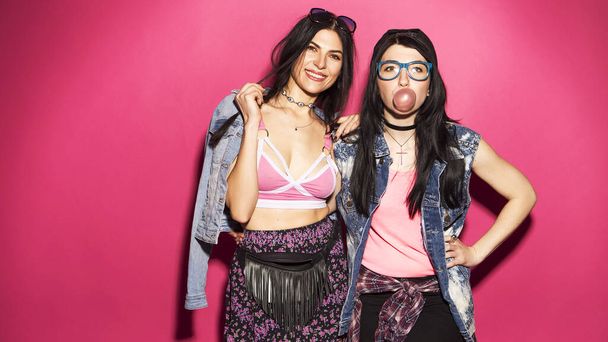 Zwei kaukasische brünette Hipsterfrauen in lässigem stylischem Jeans-Outfit haben Spaß beim Kaugummi kauen und Skateboard halten. Sie stehen auf einem leuchtend rosa Hintergrund. Fröhliche, glückliche Emotionen - Foto, Bild