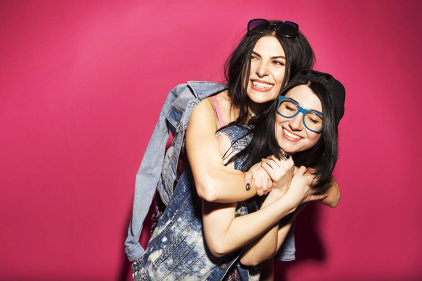 Deux femmes hipster brune caucasienne en tenue de jeans stylés décontractés s'amusent à mâcher du bubblegum et à tenir du skateboard. Ils se tiennent sur un fond rose vif. Des émotions joyeuses et heureuses
 - Photo, image