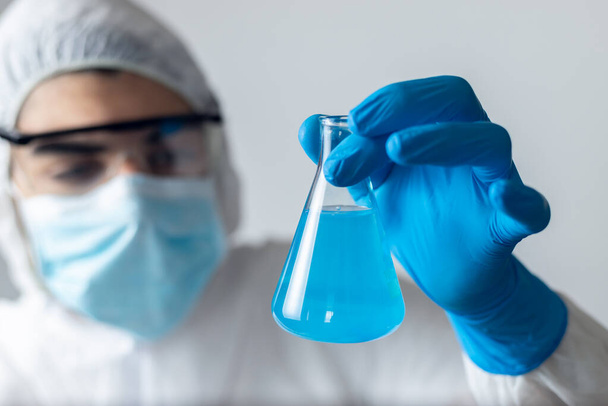 дослідник у блакитних латексних рукавичках та захисному одязі з коронавірусом, який дивиться в колбу блакитної рідини
 - Фото, зображення