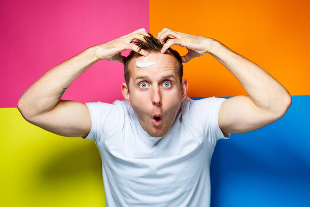 Portrait of a young, handsome, cheerful guy on a multi-colored background, dressed in a white T-shirt, creates a fashionable hairstyle, stupid and positive emotions - Photo, Image