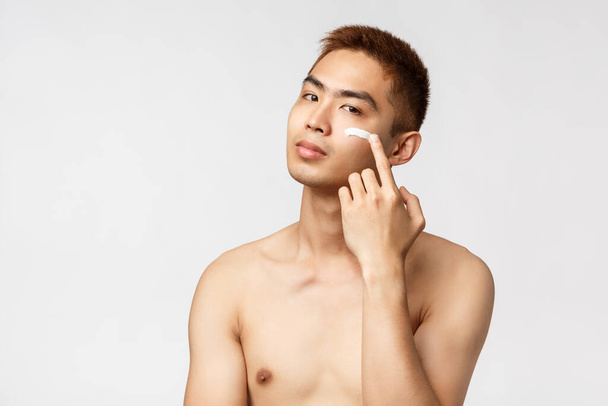 Beauty, people and leisure concept. Sassy good-looking asian man with naked torso taking care of his perfect, no blemishes skin, apply cream on face and look camera pleased, white background - Фото, изображение