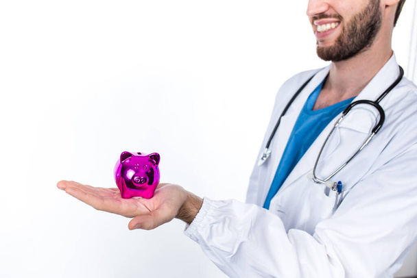 Young doctor posing holding pink piggy bank isolated on white background - Photo, Image