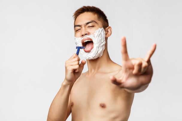 Schönheit, Menschen und Hygienekonzept. Porträt eines hübschen asiatischen nackten Mannes, der in Rasiermesser singt, während er rasiert und Spaß hat, Rock-n-Roll-Geste zeigt, Kamera wie Spiegel sieht, weißer Hintergrund stehend - Foto, Bild