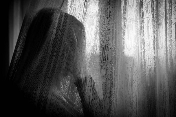 Θλιβερή γυναίκα που κοιτάζει από το παράθυρο κατά τη διάρκεια της καραντίνας λόγω ενδοοικογενειακής βίας, κοινωνικής απόστασης ή σωματικού προβλήματος - Φωτογραφία, εικόνα