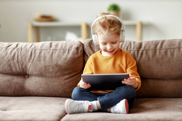 Tělo potěšený malý chlapec ve sluchátkách s úsměvem a sledování zajímavého kresleného filmu na tabletu, zatímco sedí se zkříženýma nohama na pohovce v Hom - Fotografie, Obrázek