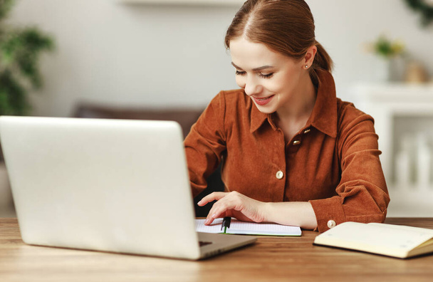 Donna allegra in camicia marrone note di lettura nel notebook e utilizzando il computer portatile mentre seduto alla scrivania e lavorare su un progetto remoto a hom
 - Foto, immagini