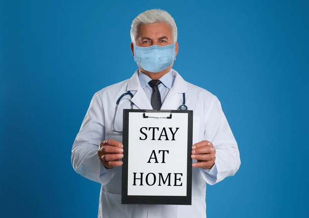 Doctor in medical mask holding clipboard with text STAY AT HOME on blue background - Fotó, kép