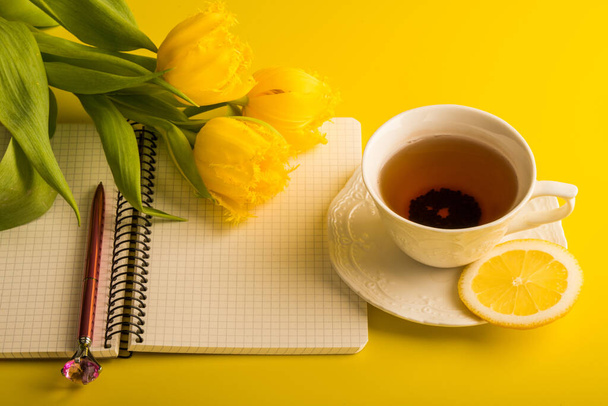 Yellow tulips flowers with cup of tea with lemon and notepad with lemon on a yellow background - Photo, Image
