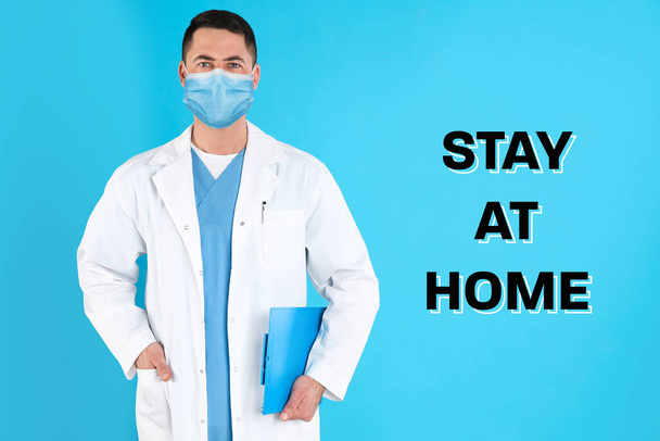 Doctor in medical mask and text STAY AT HOME on light blue background - Fotoğraf, Görsel