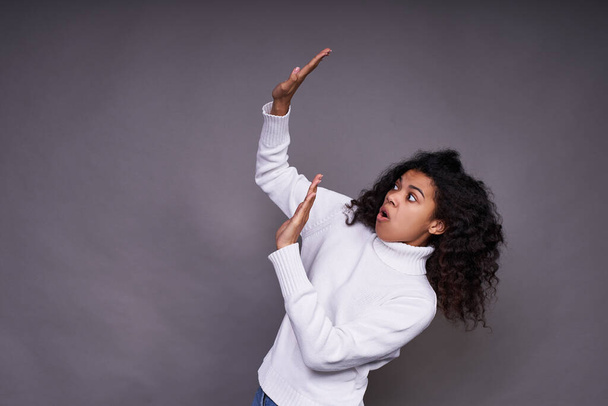 Frightened, troubled young Afro woman, in a white sweater, with curly curly hair, raised her hands up, something falls on her, she defends herself. On the left is the place to advertise your product. - Foto, immagini