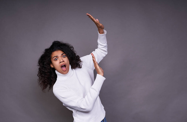 Frightened, troubled young Afro woman, in a white sweater, with curly curvy hair, raised her hands up, something falls on her, she defends herself, looks straight with her mouth open. - Photo, Image