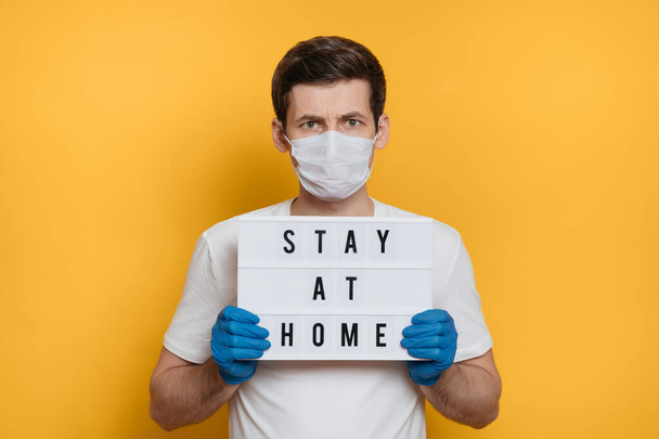 Serious man in protective mask and gloves holding lightbox with text Stay at home due to coronavirus outbreak. Covid-19 quarantine concept, pandemy. Yellow background. Stay home social media campaign - 写真・画像