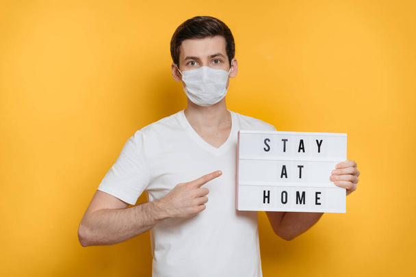 Young man in protective mask pointing with finger at Stay home sign ask people for self isolation and social distancing to prevent coronavirus infection. Yellow background. COVID-19 concept - Photo, Image