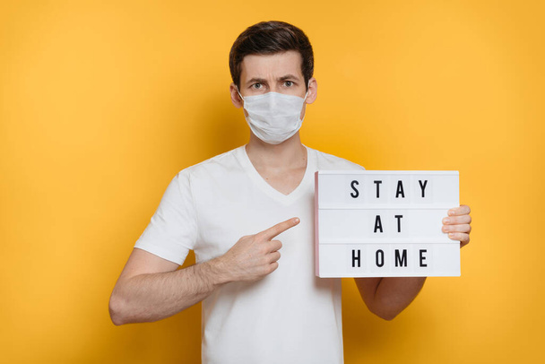 Young serious man in medical mask pointing finger at board with Stay home text. Isolated on yellow background. Stay at home social media campaign for coronavirus or COVID-19  prevention  - Fotó, kép