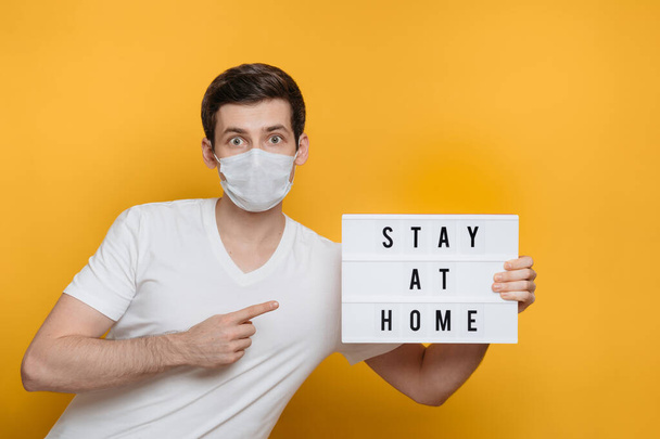 Surprised young man in face mask pointing with index finger at lightbox with Stay home text, COVID-19 quarantine concept. Yellow background. Social distancing during coronavirus quarantine. Copy space - Photo, Image