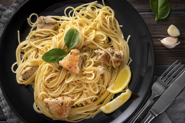 pasta  , spaghetti   with chicken fillet, lemon slices, garlic, greens, spinach,  napkin, fork, knife on a black plate on  dark ,  close up, top view - Foto, Bild