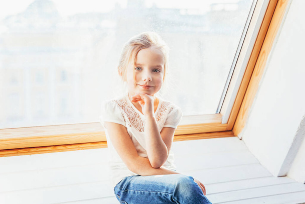 Stay Home Stay Safe. Little cute sweet smiling girl in jeans and white T-shirt sitting on window sill in bright light living room at home indoors. Childhood schoolchildren youth relax concept - Photo, Image
