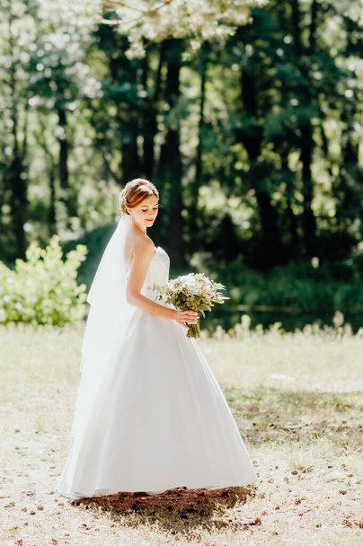 The bride in a white wedding dress and white veil is holding a bouquet of peonies in the background of a green park. Summer wedding on a sunny day. - Photo, image
