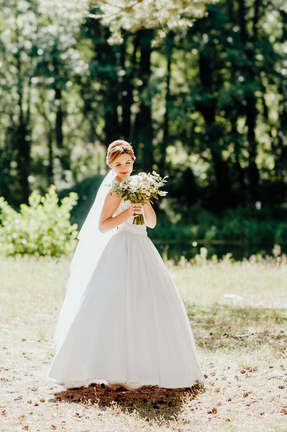 The bride in a white wedding dress and white veil is holding a bouquet of peonies in the background of a green park. Summer wedding on a sunny day. - Photo, Image
