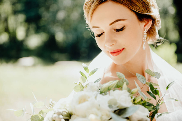 Delicate portrait of the bride on a wedding day in a snow-white dress and veil with a wedding bouquet of peonies in her hands with jewelry on her neck and ears - Foto, imagen