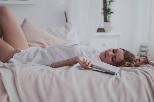 Young pretty woman in white shirt lying in a bed with a book - Photo, Image
