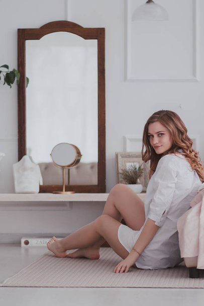 young beautiful woman in white shirt sitting on the floor near the bed and looking to the camera - Foto, imagen