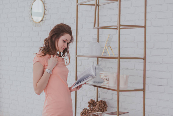 Μια νεαρή γοητευτική γυναίκα με ένα ροζ φόρεμα στέκεται κοντά σε ένα ράφι και διαβάζοντας ένα βιβλίο. - Φωτογραφία, εικόνα