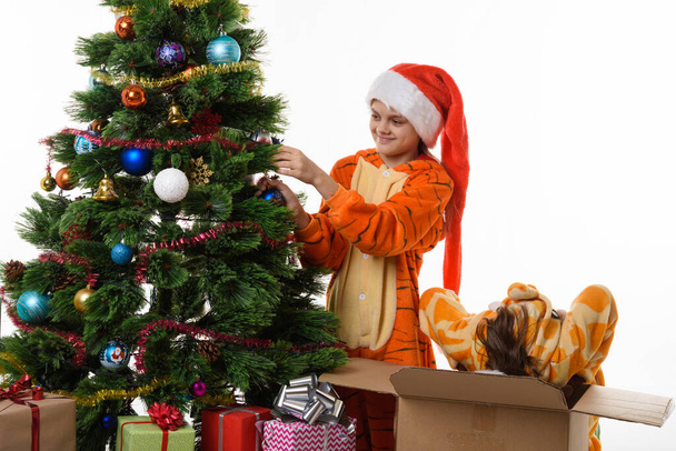 Bir kız Noel ağacına top asıyor, diğeri kafasıyla kutuya tırmanıyor. - Fotoğraf, Görsel