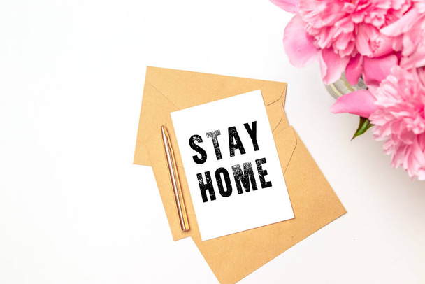 Stay home lettering made of white paper, craft envelopes, a bouquet of flowers in a vase on white table, top view. Freelance, quarantine - Photo, Image
