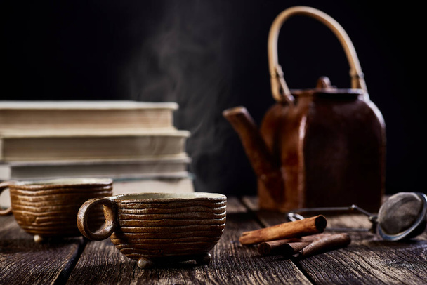 Nahaufnahme des Stilllebens mit keramischer Tasse Tee oder Kaffee, Teekanne und Zimt. Alte Holztischplatte, Rauch vor Büchern und verschwommener schwarzer Hintergrund. - Foto, Bild